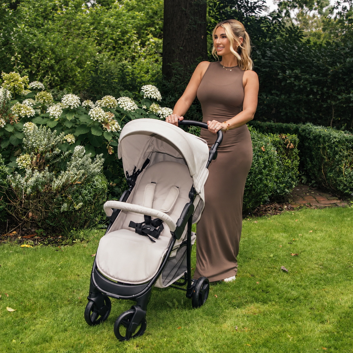 My Babiie MB30 pushchair in beige with padded reclining seat, large sun canopy and black frame, being pushed by a woman in a garden setting.