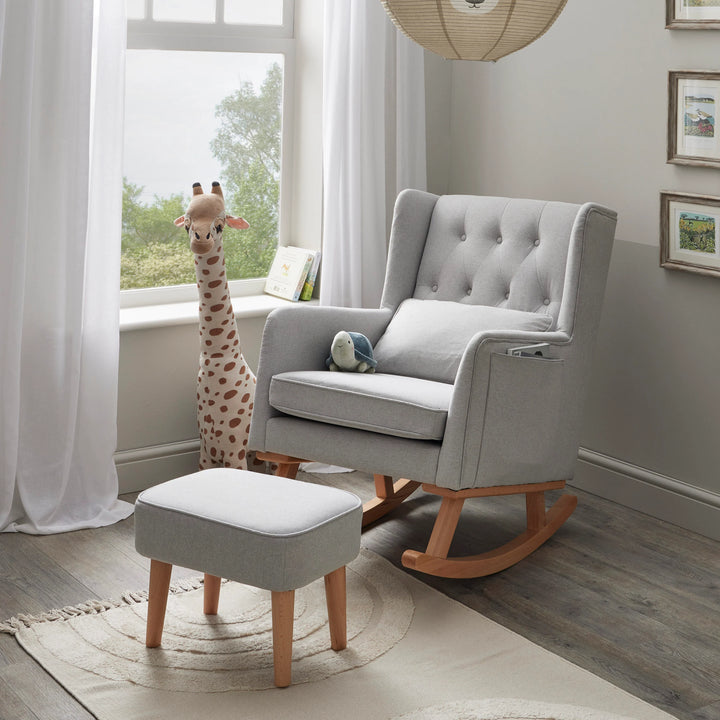 Grey rocking chair with ottoman in a room with large windows and framed pictures on the wall.