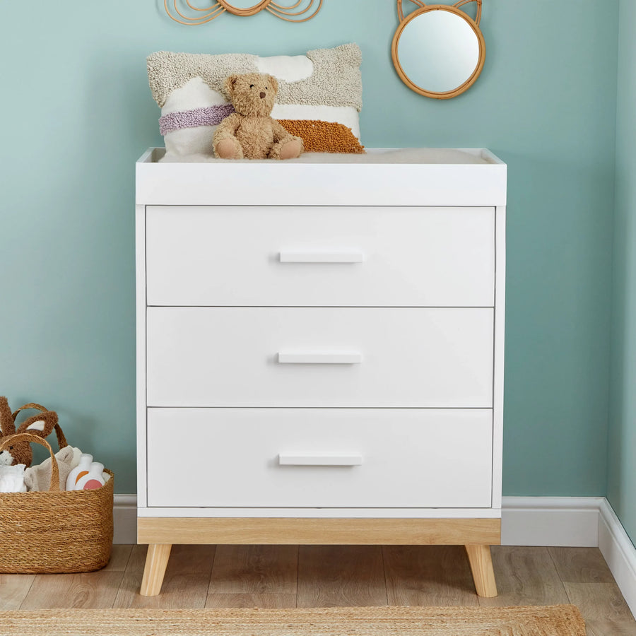 White dresser with wooden legs in a room with a teddy bear and basket on top.