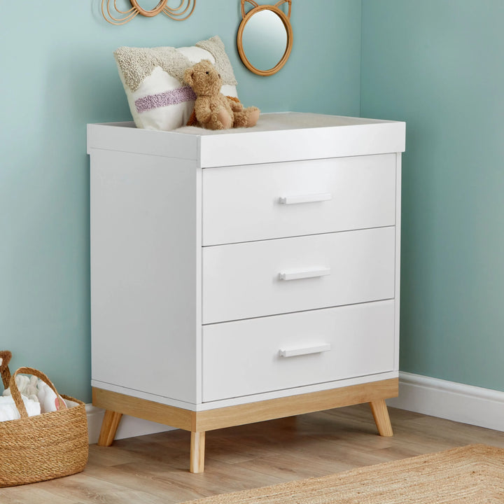White dresser with wooden legs against a light blue wall with a teddy bear and basket on top.
