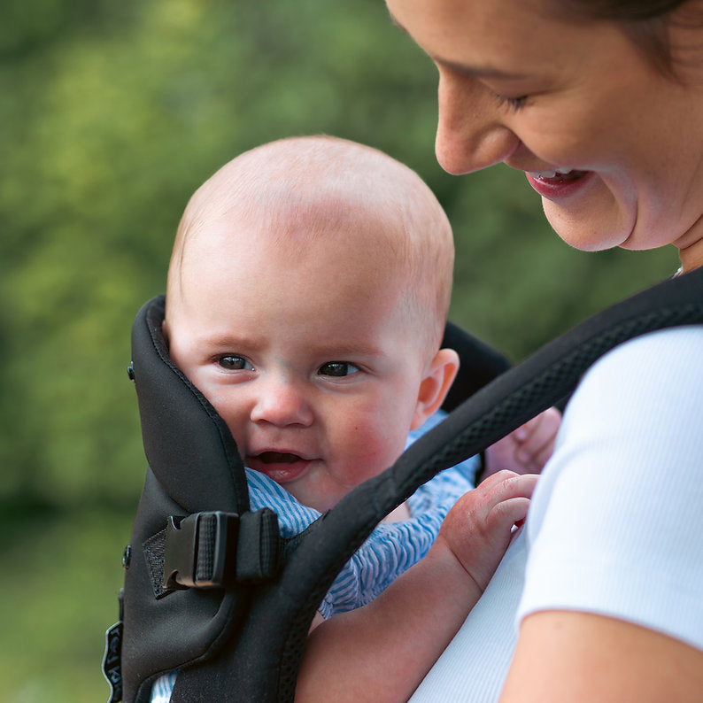 Woman holding a baby in a Red Kite Ara baby carrier outdoors with greenery in the background