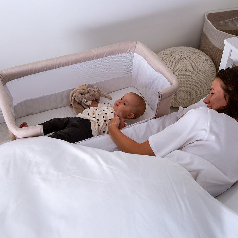 Red Kite Rockabye Bedside Crib with Baby lying in a crib next to a parent in bed, with a neutral colour scheme.